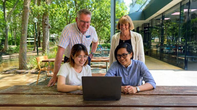 Group of university students collaborating outdoors at a campus table, discussing ideas on a laptop while working on a real-world innovation problem